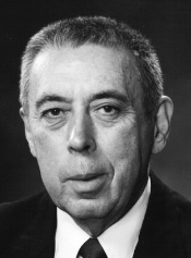 Black and white photograph of a middle-aged man with brush-cut hair, wearing a drak-colored suitcoat and necktie, and a white dress shirt