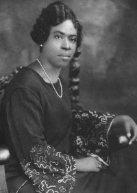 studio black and white photograph of a woman posing on a chair wearing a dress, pearl necklace, and earrings