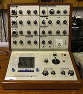 A portable L-shaped brown wooden case with a silver metal fascia filled with buttons and controls is positioned on a wooden work surface. The controls for the device are mostly rotary, and denoted with lettering and numbering. The lower part of the box contains a small matrix of holes and a joystick. Other pieces of electrical equipment are visible behind the device.