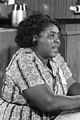 An African American woman sits at a conference, and appears to be speaking. Her hair is short and she is wearing a dress with a kaleidoscopic pattern. The picture is in black and white.