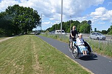 A man drives a trishaw with an elderly lady as passenger on a bicycle path along a highway in Denmark