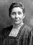 A Native American female wearing a dark shirt with a high collared white patterned chemisette. She has dark upswept hair, a solemn expression, and is facing the camera.