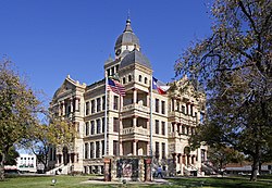 A stone building with a cloudy sky in the background. Three floors are shown with windows on each floor. There's a door entrance on the first floor and a large clock on the tower overhead.