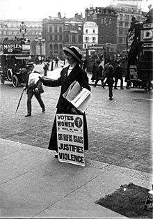 A woman hands out newspapers on a sidewalk, propping up a sign with the headline "Votes for Women"