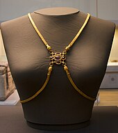 Frontal view of a grey female bust with four gold bands of many fine links, two draped over the shoulders and two plunging from the breast bone sweeping below the breasts and going behind the back. The four converge between the breasts, where each band ends in a head which connects to a centrepiece, a purple gem surrounded by eight smaller sockets, four empty and four with red stones