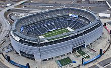 Aerial image of a large American football stadium with three tiers of stands and no roof; the stands are empty and several pieces of equipment are parked around its exterior.