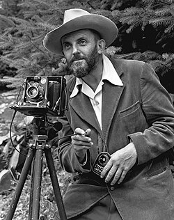 A photo of a bearded Ansel Adams with a camera on a tripod and a light meter in his hand. The photo was taken in 1950. Adams is wearing a dark jacket and a white shirt, and the shirt collar is spread over the lapel of his jacket. He is holding a cable release for the camera, and there is a rocky hillside behind him.