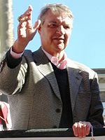 A gray-haired man stands in the back of a truck. He is wearing a patterned jacket, a pink shirt and a dark sweater vest. His right hand is raised.