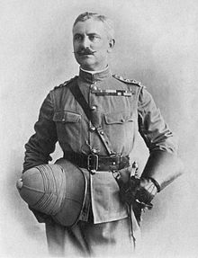 Mustached man in officers uniform. With San-Browne belt, sword at waist, wearing gauntlets and holding his pith helmet at waist level in his right hand.