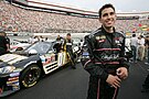 A Hispanic racecar driver dressed in full racing gear poses besides a black race car.