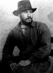 Black-and-white photo of Johnson wearing a hat and sitting on a chair with his hands on his lap.