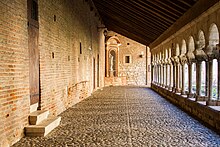 Photo couleur d'une galerie de cloitre entre un mur de briques à gauche et une colonnade à arc roman à droite, donnant sur le jardin. Le sol est pavé de galets et la toiture simple penche vers le jardin.