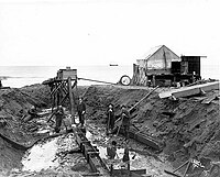 miniature lift at beach, 1904, showing four men with showels in a hole on the beach with the sea in the background