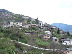 Image of the hillside village of Kosovo, taken from the neighboring hillside. A mixture of about 50 old and new houses, a few have partially collapsed. The hillside is heavily forested, the only visible road is narrow, and snakes gently through the valley below.