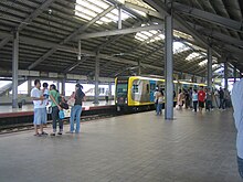 A relatively empty boarding platform with only a handful of people identified by a sign as that at Vito Cruz station. Pebbles line the tracks and sunlight comes in from spaces open to the outside and large open flaps in the dark warehouse-like roof.