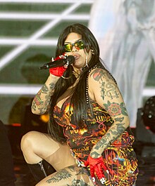 The singer Tomasa del Real wearing a pair of hoop earrings, dress with fire printed on it, red gloves and black knee length boots kneeling down on her left knee holding up a microphone to her mouth with her right hand.