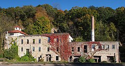 A wide stone building with two- and three-story sections, a smokestack, and ivy, with a forested bluff behind