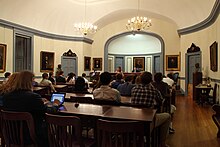 A collection of people sitting in a white room that has paintings on its walls.