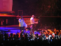 In a stage, two men, both wearing white shirts, can be seen holding microphones. The first man, wearing a long sleeve shirt, sings. While the other man, wearing a shirt, dances to the music played. A crowd of people can also be seen holding up their cellphones.