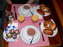 Table set with mugs and glasses of brown liquid, carafe, plates of pastries, butter, and tiny jams.