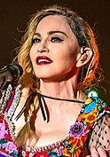 A woman with plaited hair, blue eyes and red lipstick wearing a colorful dress and guitar strap.