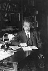 Einstein, sitting at a table, looks up from the papers he is reading and into the camera.