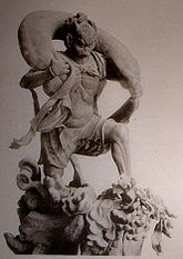 Three-quarter view of a statue. His left leg is bend as if climbing stairs and he is carrying a long bag-shaped object which goes from one shoulder to the other around the back of his head. Black and white photograph.