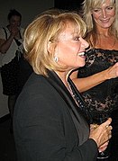 A smiling blonde woman wearing a light blue blouse and an elaborate necklace stares at the camera.