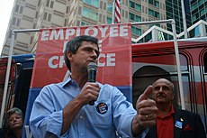 A gray-haired man wearing a blue shirt and black campaign button speaks into a microphone at an outside political rally. A man and woman stand behind him, and a banner reading "Employee Free Choice" can be seen in the background.