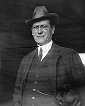 Black and white photo of a white man in a three-piece suit with glasses and a hat.