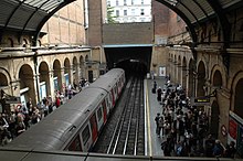 A train sits in a station in a cutting with platforms each side. The retaining walls of the cutting feature a series of tall buttresses linked with arches at the top. A glass roof arches above the tracks and platforms.