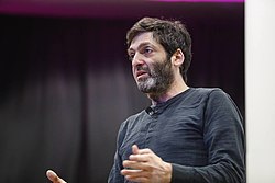 Dan Ariely wearing a dark long-sleeved shirt, standing in semi-profile, appearing to speak onstage