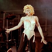 Madonna wearing a beige corset and black pants. She has blonde curly hair and has a headset microphone to her mouth.