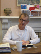 grey haired man in white shirt with glasses seated at table reading from a book