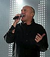 A man with a shaved head wearing black and singing into a microphone. The background contains dotted lighting on a stage.