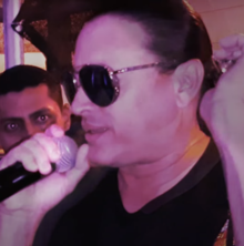 A man wearing sunglasses is facing left holding a microphone.