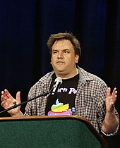 A brown-haired man with a light brown plaid shirt stands behind a podium against a black background.