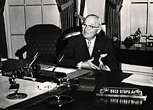 Man in suit sitting behind desk with sign that says "The buck stops here"