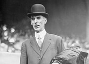 A man standing in a baseball stadium wearing a suit and bowler hat. He has a coat folded over his left arm.