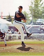 A man in a blue baseball jersey, a blue hat, and white pants stands on a pitcher's mound and prepares to throw a pitch.