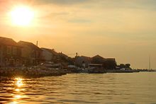 A photograph of a coastline with a bright sun in the sky above that reflects on the water below and darkens the buildings on the coast