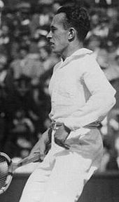 A black-haired man all dressed in white, holding a wooden racket in his right hand