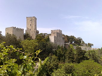 A castle with a tall narrow tower and walls topped by battlements stretches along the edge of a cliff covered in trees and palm trees.