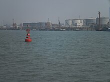 A red port-side buoy with an oil wharf and tankers in the background