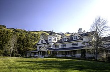 A large white house with black roofing in front of green hills and forests.