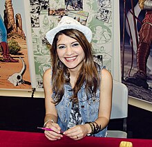 Portrait photo of a seated young woman with wavy brown hair who is wearing a white hat and blue jeans vest, and is smiling at the camera, while holding a pink pen.