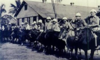 A black-and-white photo of a series of men in uniform riding bulls in front of a building