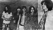 Five smiling men in a row, diagonal to camera angle. The man on the left (farthest to the back) has very long hair and a full beard; he wears a white T-shirt and tie-dyed pants. Next to him, Dave Davies, also with very long hair, wears reflective sunglasses, a black short-sleeved shirt and jeans. In the middle, Mick Avory wears an unbuttoned leather vest and white pants. The man to his right wears a heavy, probably brown leather jacket with a design that is possibly Native American. On the far right, in front, Ray Davies wears a giant paisley kerchief knotted like a tie over a white jacket.