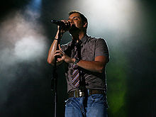 A man with a gray buttoned shirt with a tie is performing on stage.