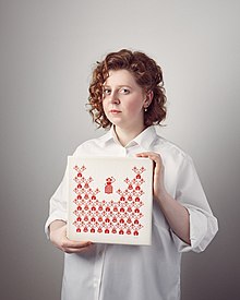 Rufina Bazlova is depicted standing. She is holding a square artwork with red pictograms on a white background. Bazlova's red curly hair is half-length and she is wearing a loose white blouse. She looks directly into the camera.